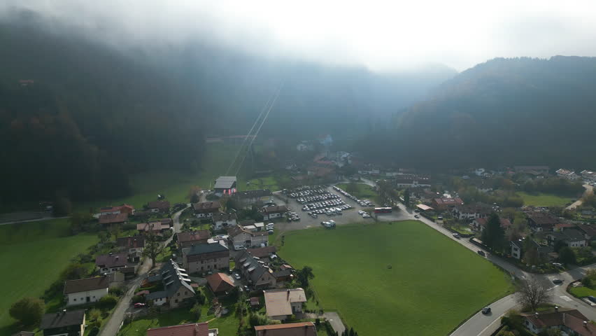 Scenic aerial shot of Bergen, a picturesque Bavarian village with clustered homes, green fields, and a cable car station, nestled below mist-covered mountains under a soft, diffused sky.