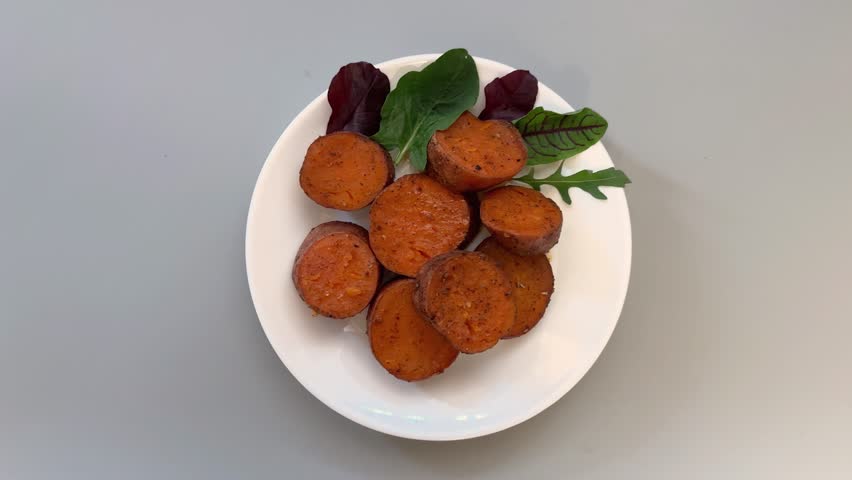 Portion of the baked circle slices of the sweet potato tubers in their skin served with greens leaves on the dish on a gray background, top view while approach with rotation
