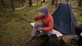 Male hiker drinking tea and surfing Internet on smartphone in tent camp during hiking in fall in woods. Backpacker makes route in map in phone and drinks hot drink from cup in background of tent.  - Powered by Shutterstock - Get 15% off with code: PIKWIZARD15