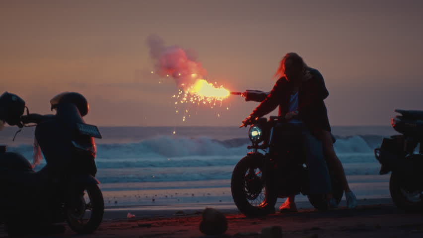 Romantic woman and man ride on moto bike, Bali Indonesia asia, twilling sunrise. Two young couples riding on vintage motorcycles with red burning signal flares after sunset on beach, slow motion fire