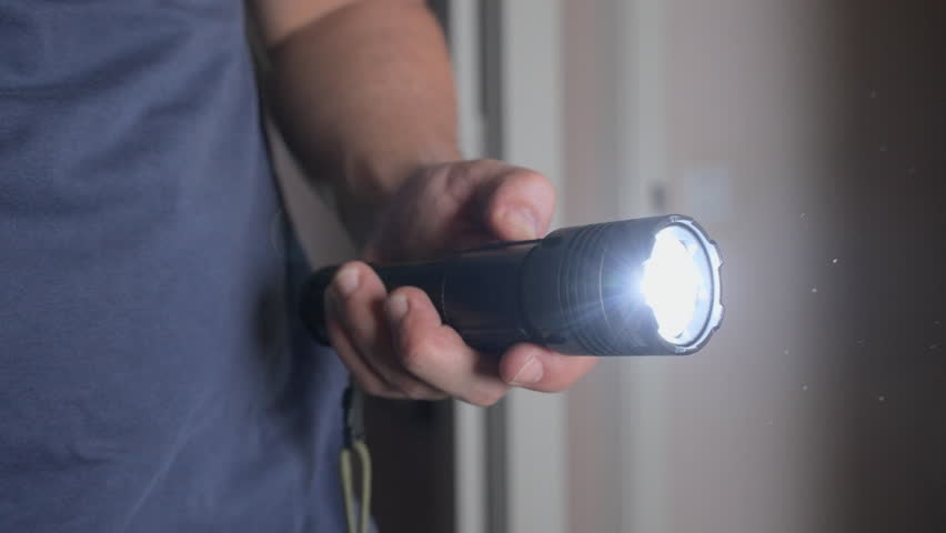 A person’s hand turns on a flashlight in a dark room. The beam of light cuts through the darkness, creating a mood of tension or exploration