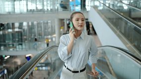 A asian businesswoman exudes confidence and happiness as she navigates an escalator while chatting on her mobile phone in a vibrant airport lobby, enjoying a networking moment. - Powered by Shutterstock - Get 15% off with code: PIKWIZARD15