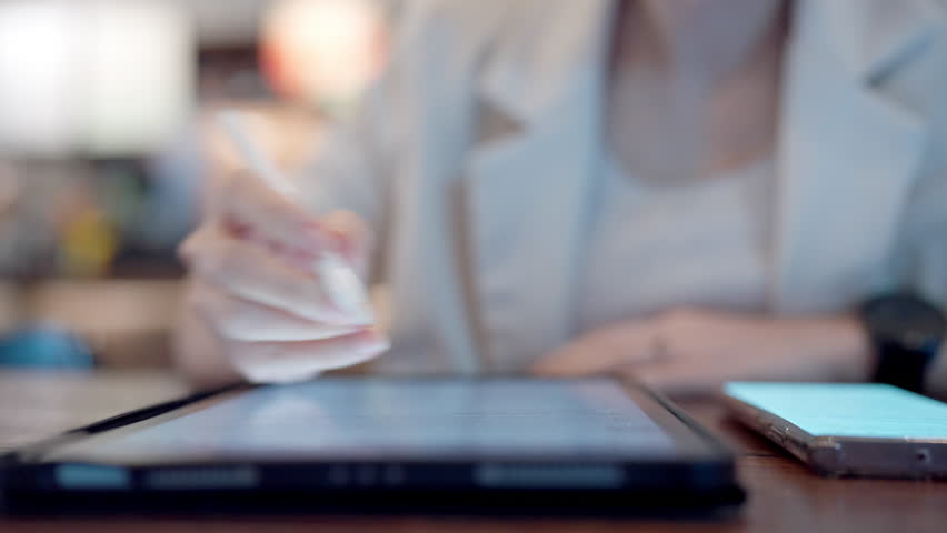 A woman is writing on a tablet with a pen. A cell phone is on the table next to the tablet