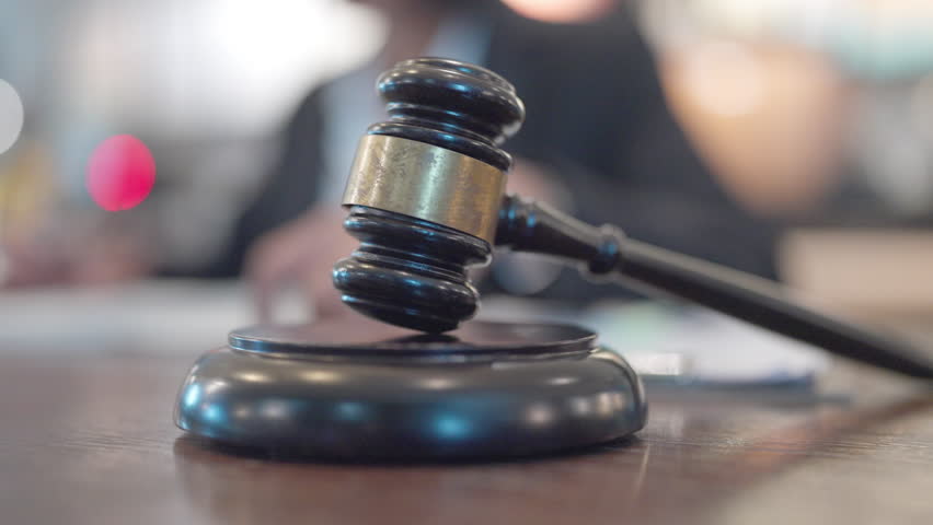 A black gavel sits on a wooden surface. A person is sitting behind the gavel. A person is typing on a keyboard