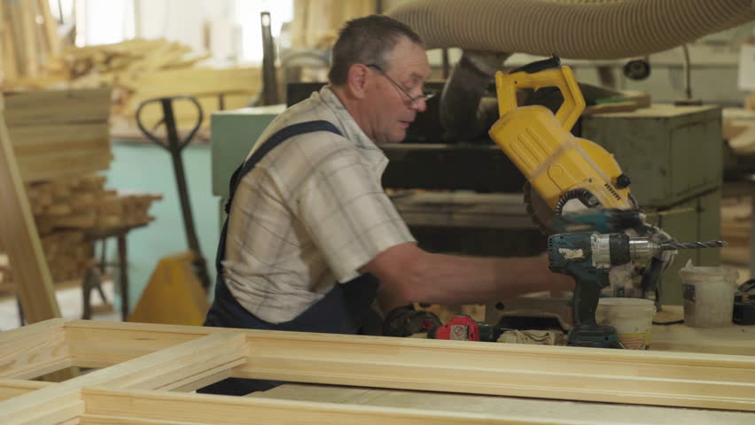 Joiner Picks Wooden Plank As Material For Production Of Window Frame. Joiner Puts Down Electric Drill Tool. Crafting Frames From Timber Planks In Workshop. Joiner Uses Planks For Woodwork