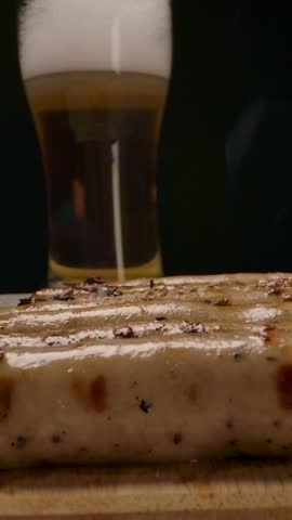 Pan around view of bunch of steamy grilled sausages placed on wooden board near cup of fresh beer in dark room. Vertical shot.