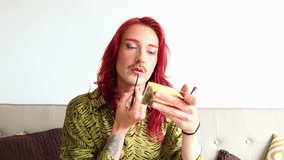 Non-binary model with long red hair and tattoos applying lipstick while looking in a small golden mirror, sitting on a beige sofa, celebrating body positivity and gender expression - Powered by Shutterstock - Get 15% off with code: PIKWIZARD15