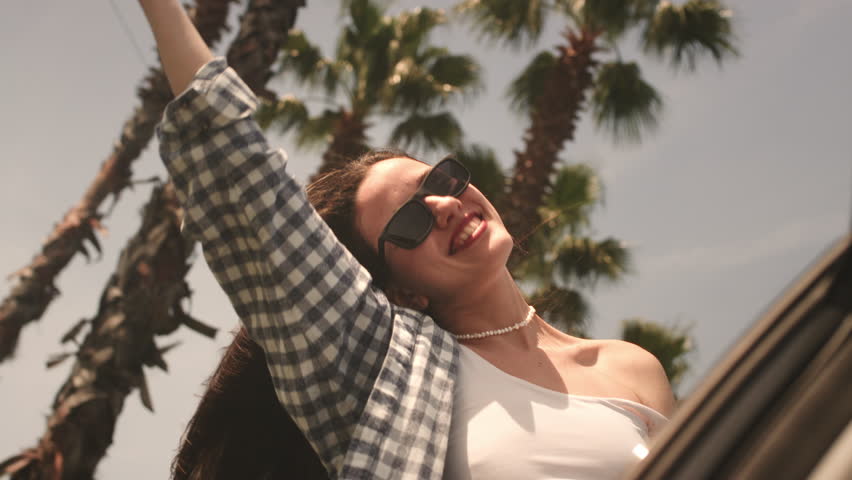 Excited woman in the window of a rental car, hair moving in wind. Caucasian female is happy to see palm trees by sea on beach. Hipster girl in shirt makes waves with her hand, goes surfing