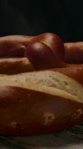 Pan around view of hot fresh pretzel emitting stream while placed on wooden table in dark room. Vertical shot.