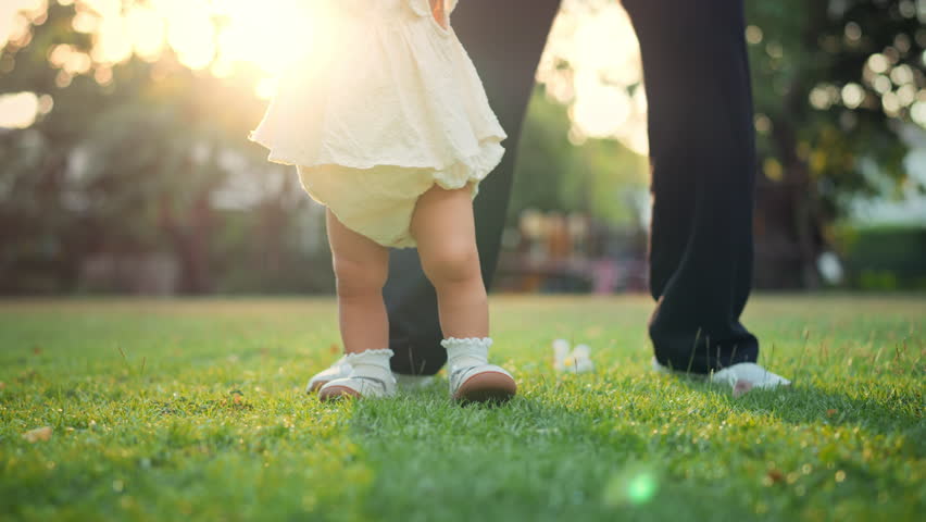 Close-up of baby wearing shoes taking first steps on green grass in park with soft morning sunlight – toddler walking with mother, childhood milestone and outdoor lifestyle concept