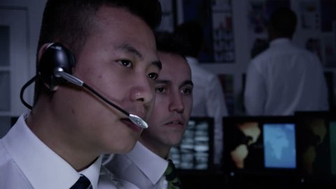 A team of security personnel man the stations within a busy system control room. Could be a weather or power station or air traffic control. It could be a  police/army control facility. Slow motion.