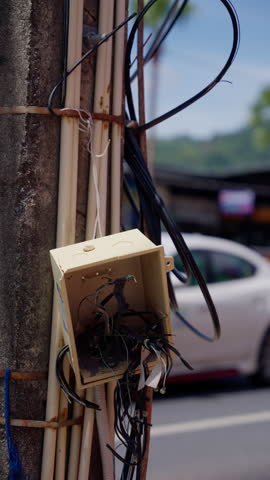 Exposed electrical wires hanging loosely from junction box near roadside pole, thailand urban street blurred background