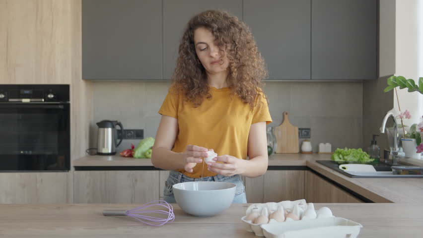 Young Woman Cracking Eggs Into Bowl While Preparing Breakfast In Modern Kitchen