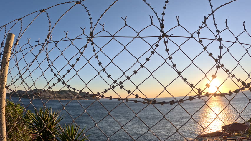 Border with barbed wire at sunset. Sunset casting across razor wire fence, revealing stark contrast between serene ocean landscape and restrictive boundary, symbolizing freedom versus confinement