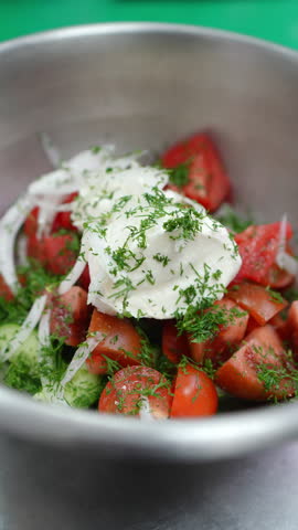 Vertical footage Professional chef combining fresh summer vegetables with creamy dressing, mixing tomatoes, cucumbers, onions, and dill in metal bowl for vibrant vegetarian salad preparation