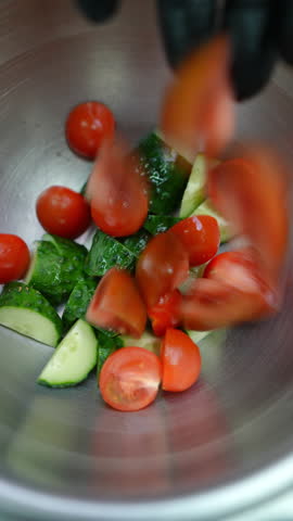 Vertical footage Chopped red tomatoes cascading into metallic bowl with green cucumbers and small cherry tomatoes during professional food preparation in commercial kitchen setting