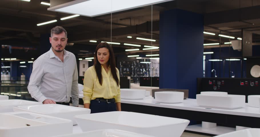 Young happy couple choosing sink for bathroom in their house standing at building market or shopping mall planning repair. Man and woman in bath store looking to showcase of plumbing shop. 4k video.