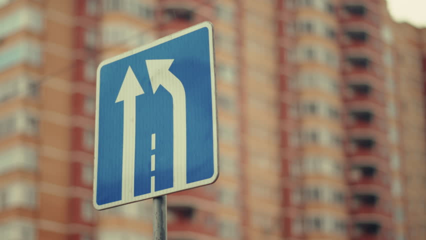 Slow motion capturing road sign showing lane directions, blurred urban buildings rising behind, emphasizing traffic guidance and metropolitan infrastructure