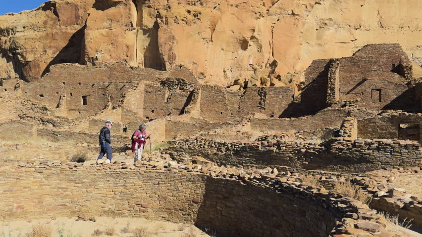 Some tourists walking among large stone ruins of a desert cliffside archaeological site in Chaco Canyon, New Mexico.