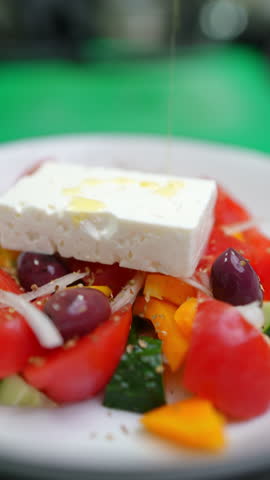 Vertical footage Feta cheese is placed on a traditional greek salad with tomatoes, cucumbers, onions, olives, peppers and olive oil being poured on top