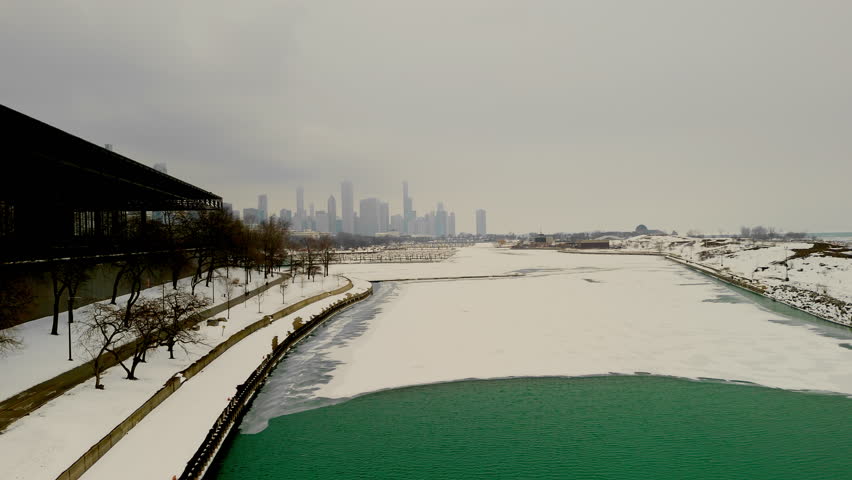 The aerial view of snowy marina on urban Chicago skyline. Frozen harbor, towering buildings in the background. Empty docks with patches of icy green water through the snow