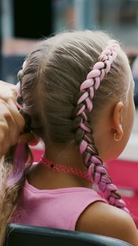 Beautiful blond girl, of seven years old, braided two pigtails, do a hairstyle with pink locks of hair in a beauty salon, a hairdresser