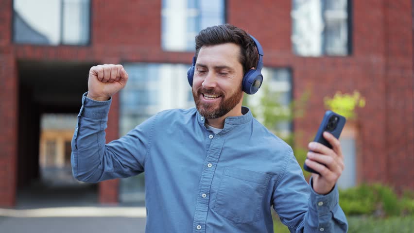 Positive man in casual clothes enjoys music through wireless headphones in city. Male feels happiness, joy, and freedom.