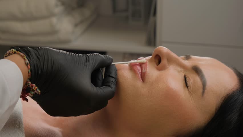Aesthetician applies numbing cream to a woman's lips with a cotton swab before a cosmetic injection procedure. The woman is lying on a treatment bed in a beauty clinic.