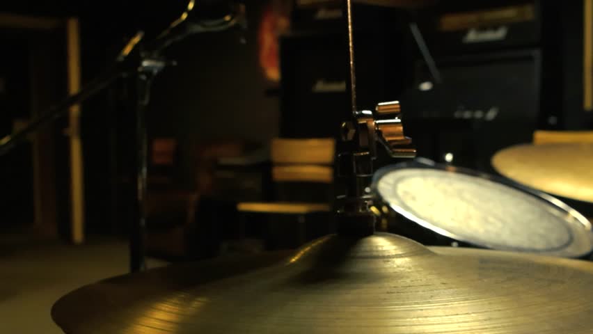 Rotation around an acoustic drum set and cymbals in a dark studio room, where small lights cast a soft glow on the drum kit. The interplay of light and shadow highlights the textures of the cymbals