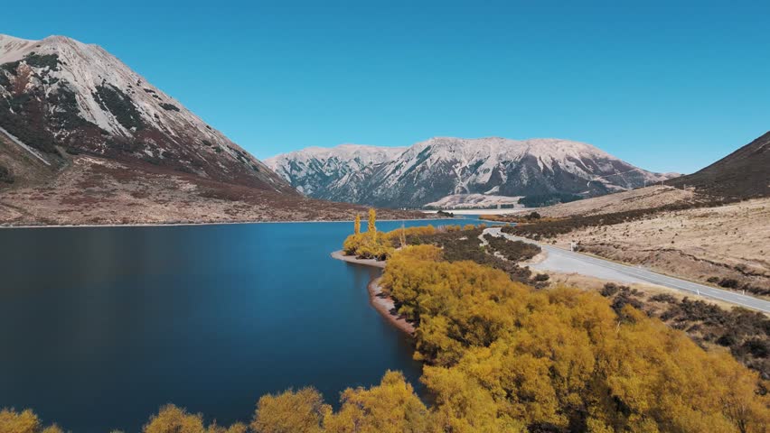 Drone pull-out reveals Lake Pearson alongside State Highway 73 and Purple Hill in Arthur’s Pass, New Zealand, captured on a sunny autumn day with vibrant colors and clear alpine scenery.