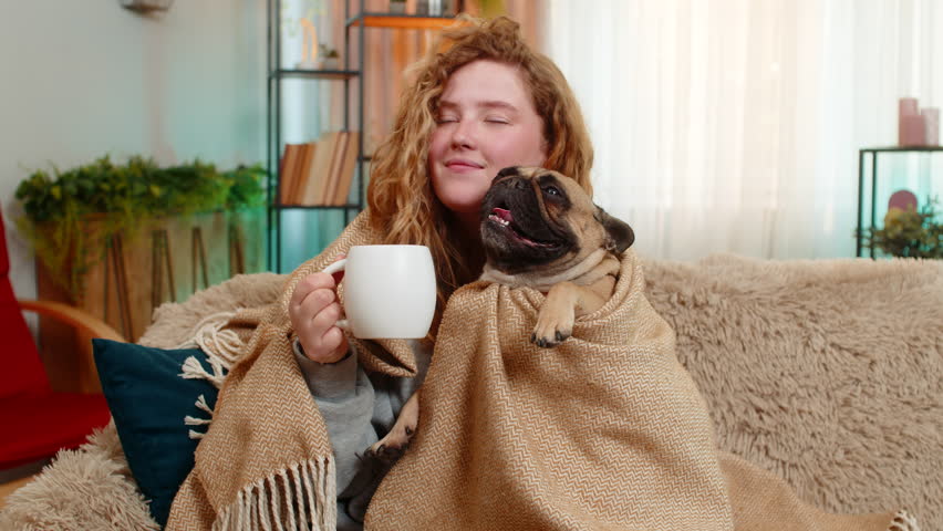 Young Caucasian redhead woman enjoys tea under blanket on home sofa, holding cup with warmth and calm. Pug dog rests beside her under same blanket, sharing comfort and peaceful happy family moment.
