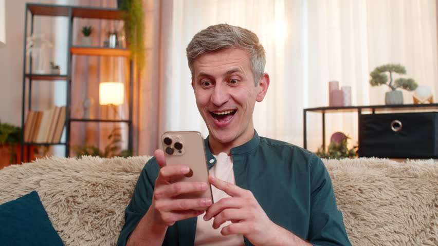 Young man at home reacts on sofa with wide eyes and open mouth to great wow message on smartphone. Caucasian guy shows happiness and surprise after seeing exciting update, offer, or good opportunity