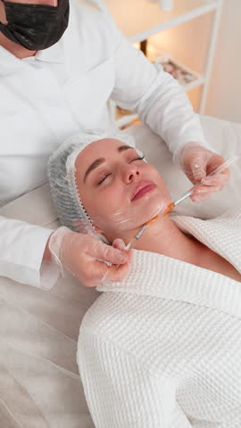Enzyme therapy. Beautician applying moisturizing medical peeling cream mask on young adult woman face with brushes. Girl skin care procedure on spa. Facial cosmetology treatment. Vertical video shot