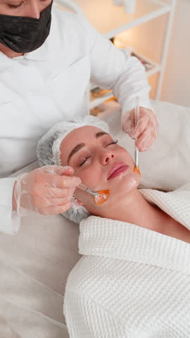 Enzyme therapy. Beautician applying moisturizing medical peeling cream mask on young adult woman face with brushes. Girl skin care procedure on spa. Facial cosmetology treatment. Vertical video shot