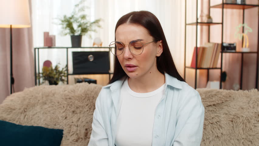 Young woman at home feeling eye pain and discomfort, tiredly removing glasses and rubbing eyes gently. Caucasian sick girl on sofa experiencing visual strain, fatigue from screen work or reading