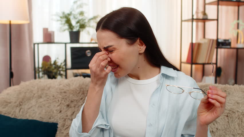 Young woman at home feeling eye pain and discomfort, tiredly removing glasses and rubbing eyes gently. Caucasian sick girl on sofa experiencing visual strain, fatigue from screen work or reading