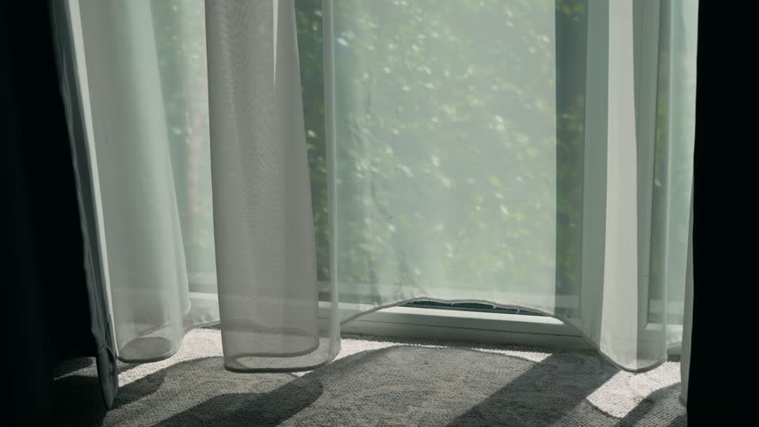 Wind blowing in open window and swaying thin curtain in living room. Breeze flowing through open glass and fluttering sheer drape inside cozy suite. Air moving through wide window and waving light
