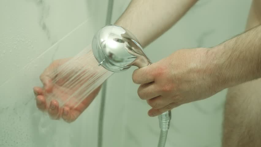 Man testing water temperature with hand under shower stream. Holding showerhead while preparing for bath. Adjusting water flow in modern bathroom. Morning hygiene routine in progress with careful hand