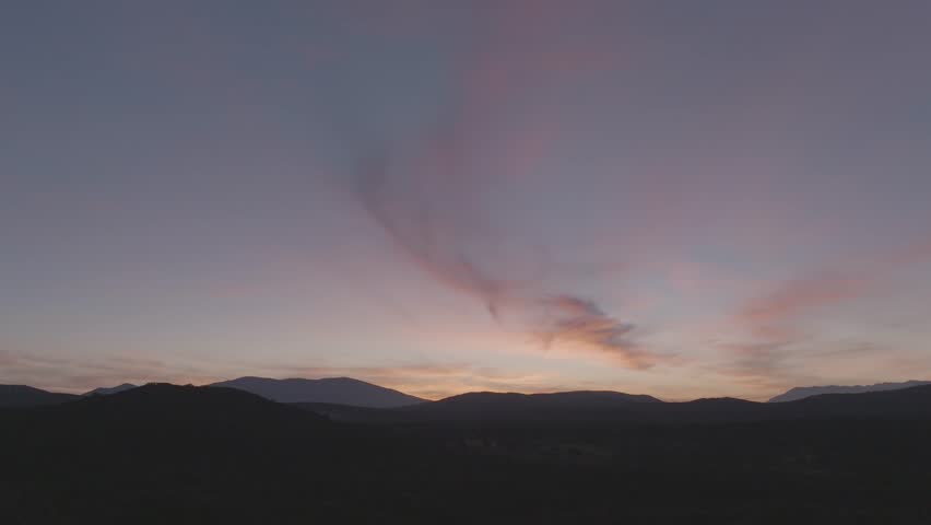 Aerial drone shot at dusk in 70mm showing dark mountains and a mostly clear blue sky, with a faint, fading cloud across the middle. Camera rises to leave only the open sky in frame.