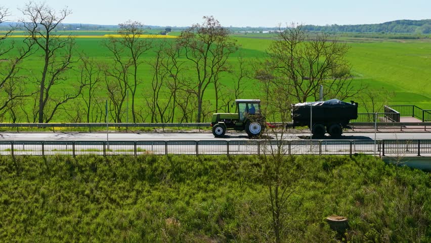 Farming tractor driving on road with trailer and trees in background. Tractor driving along countryside route passing fields and tree line. Agricultural tractor driving through rural area pulling