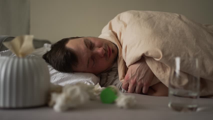 Caucasian man suffering from cold or flu lying in bed covered with blanket, checking his body temperature with a thermometer, blowing his nose and coughing, with tissues and glass of water near him