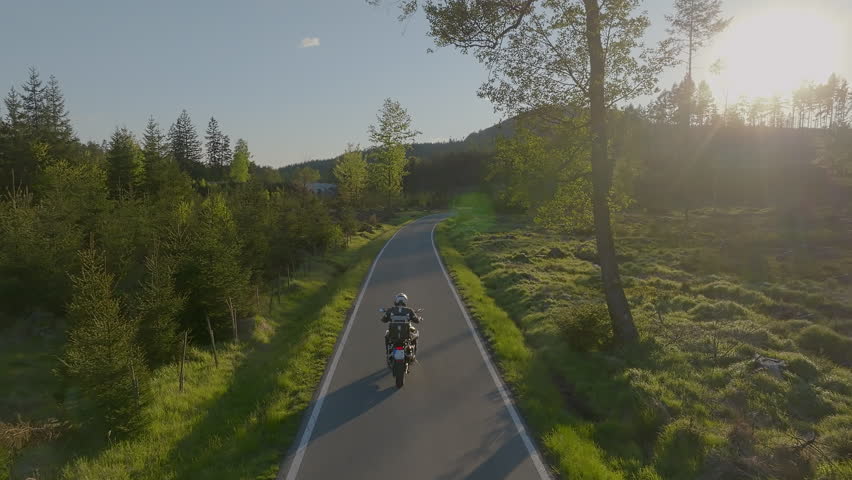 Driver riding motorcycle on empty asphalt road, spring mountains during sunset