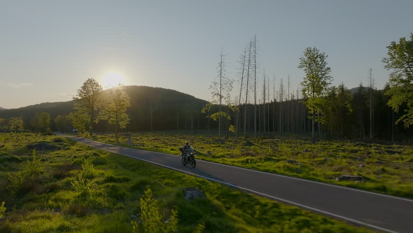 Driver riding motorcycle on empty asphalt road, spring mountains during sunset