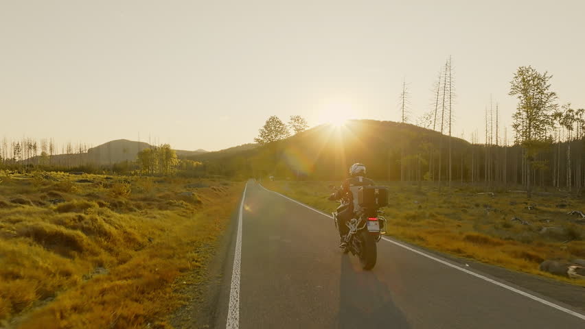 Driver riding motorcycle on empty asphalt road, spring mountains during sunset