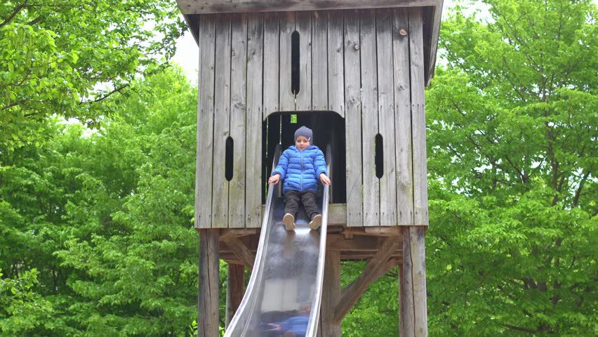 Child in blue jacket sliding down a wooden playground slide. Surrounded by lush green trees, the child displays a joyful expression, experiencing outdoor fun and play.
