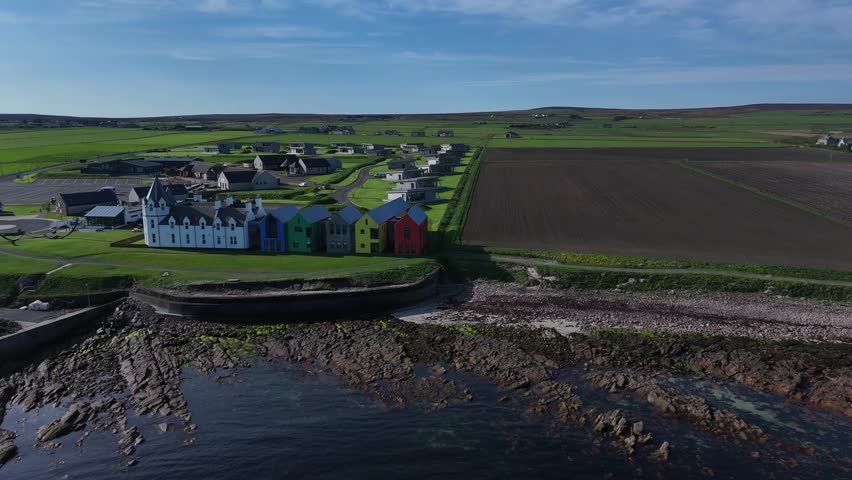 Panning shot of John O'Groats harbour with colourful buildings next to the signpost