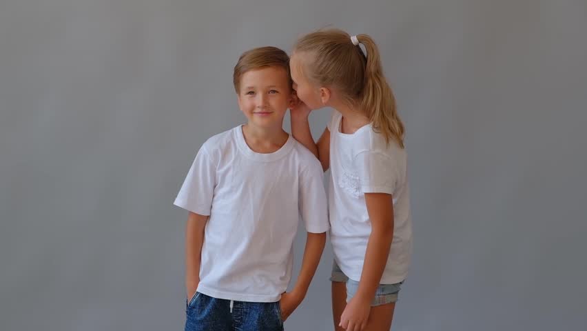 Two children, a girl whispering into a boy's ear against a plain grey background. Suggests sharing a secret, telling a story, or a moment of intimacy between siblings or friends