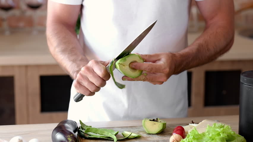 Young Man Peeling Ripe Avocado In The Kitchen, Preparing Healthy Vegetarian Breakfast Ingredients