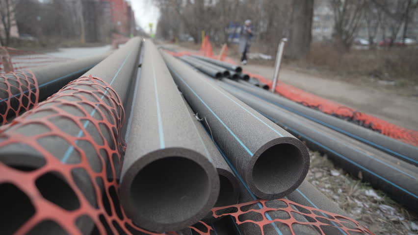 Thick rubber pipes rest on cold ground awaiting installation. Industrial materials lie in construction zone. Durable tubes remain idle