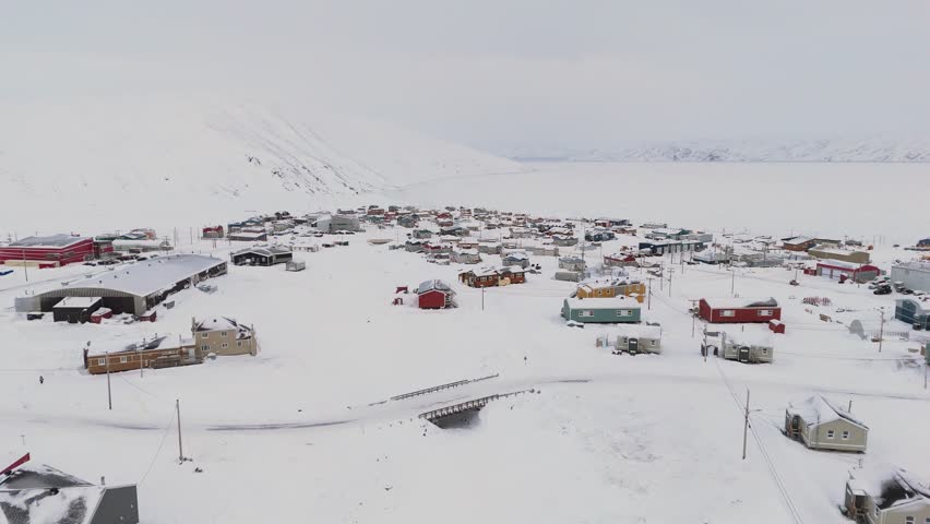 Drone footage of a snow-covered Arctic town surrounded by vast tundra and distant mountains. The shot showcases colorful structures of a northern Canadian community under overcast skies.